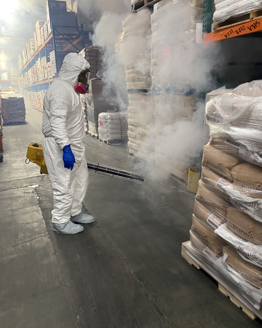 Raknid technician in full Tyvek coveralls and respirator applying a ULV treatment along a pallet racking aisle in a food ingredient warehouse