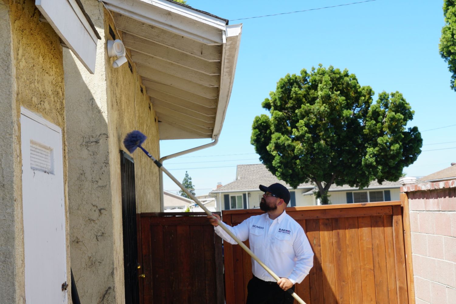 Ricardo Ayala in a Raknid Pest Solutions uniform shirt de-webbing the eaves of a property with an extension brush